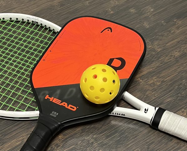 The bright orange design of a pickleball racket pops out on a court, supported by a traditional tennis racket.